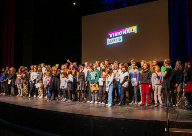 Gruppenfoto (klein) VL22 Foto der Bühne mit allen Nominierten der VISIONALE LEIPZIG 2022, im Hintergrund ist das Logo der VISIONALE zu sehen.